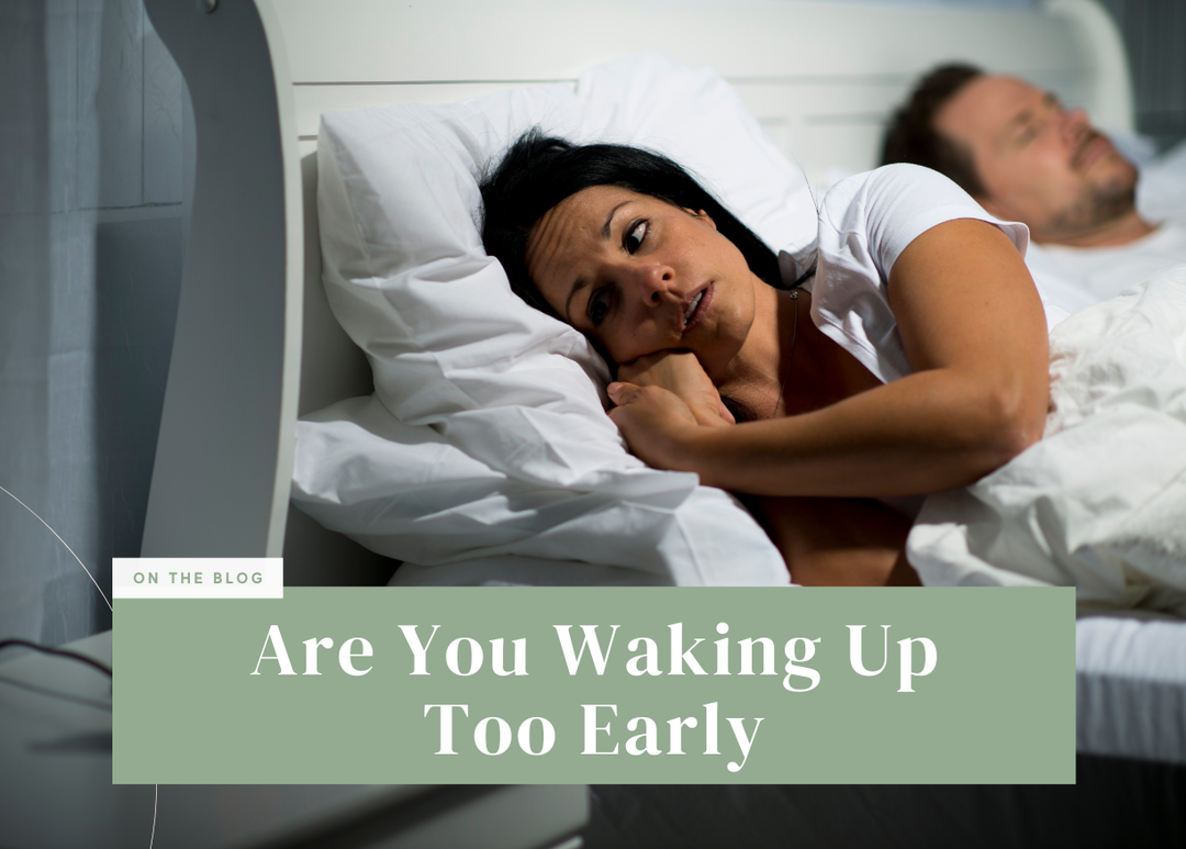 Dark-haired woman lying awake in bed with a concerned expression while her partner sleeps nearby.